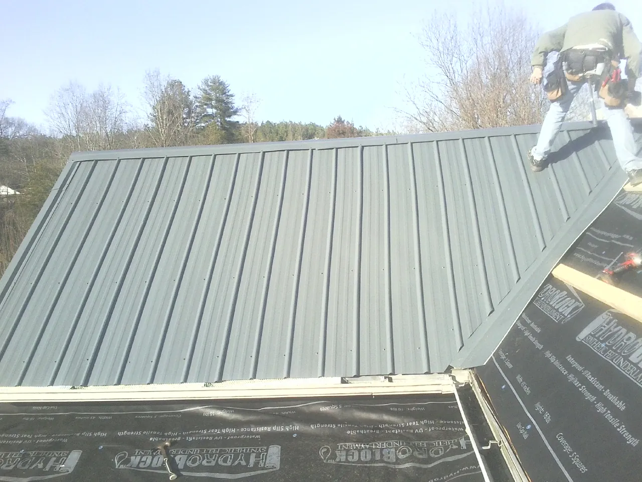 Crew installing dark green standing seam panels over synthetic underlayment in Watertown